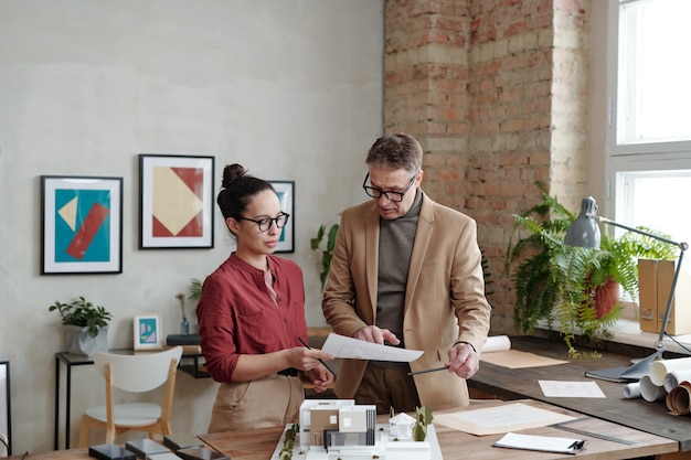 Chief architect in jacket standing at desk with 3D model of cottage house and giving recommendation to young employee according to sketch