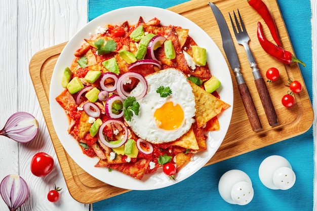 Chilaquiles with a fried egg, ham, crumbled panela cheese, avocado and tomato salsa on a white plate on a  cutting board, Mexican dish, view from above, close-up