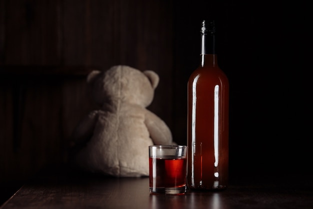 Child abuse concept. Teddy bear as a symbol of child's safety and bottle with glass in a dark room.