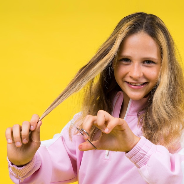 Child adorable girl hairdresser cutting long blonde hair with metallic scissors on yellow