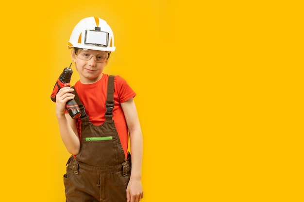 Child as builder Boy in white helmet protective glasses wears work jumpsuit and holds drill Isolated on yellow background