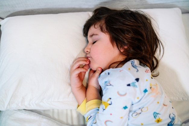 Child asleep in bed with closed eyes and innocent face, wearing pajamas and white sheets