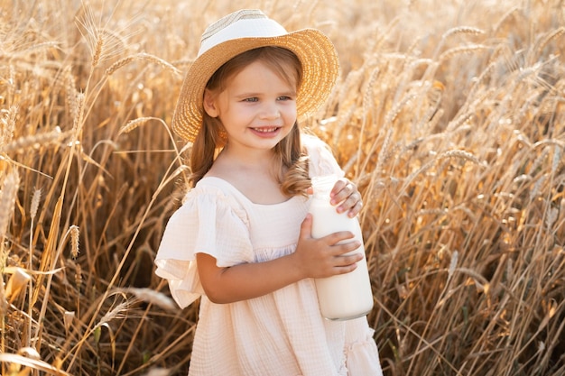 child blonde girl in straw hat and beige muslin dress holds bottle of milk in wheat field on sunset