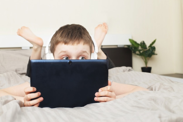 Child boy lying on bed at home covered his face with a pc tablet with wireless headphones