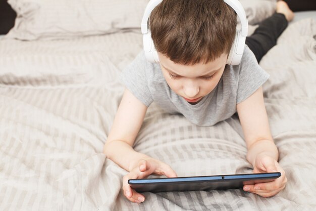 Child boy lying on bed at home with pc tablet  with wireless headphones