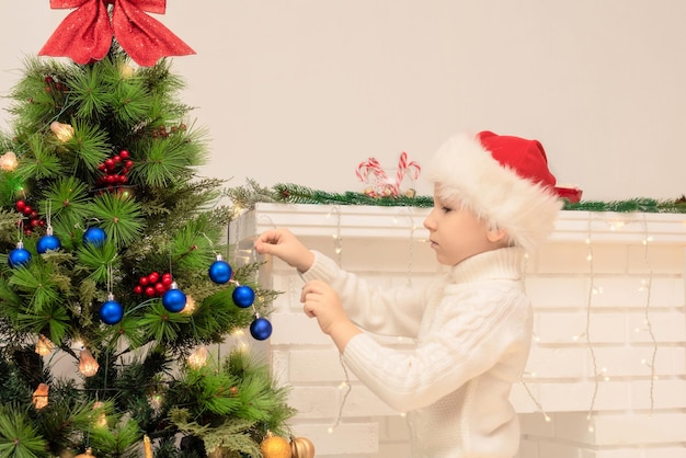 Child boy in red Santa hat decorates Christmas tree close up Xmas or New Year cozy holidays concept Selective focus