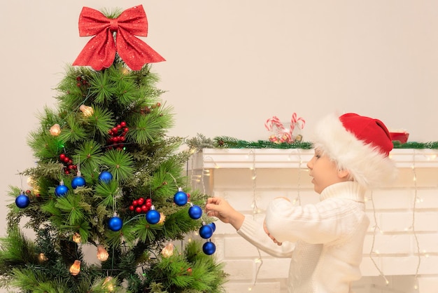 Child boy in red Santa hat decorates Christmas tree close up Xmas or New Year cozy holidays concept Selective focus