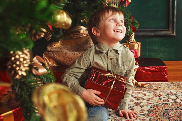 Child boy sitting under the Christmas tree with gift box.