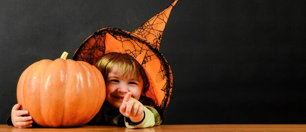 Child boy in witch hat with halloween pumpkin pointing to you trick or treat happy holidays