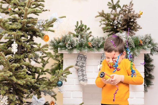 Child boy with petard in his hands near decorated Christmas tree and fireplace Winter or New Year cozy holidays concept Xmas background with selective focus