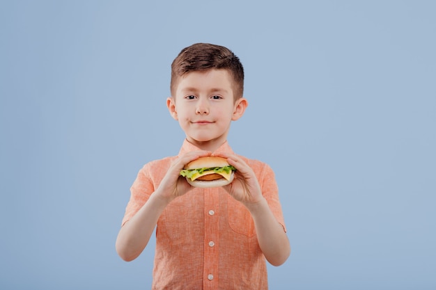 Child boy with sandwich in hand looks at the camera isolated on blue background copy space