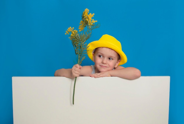 Child in bright yellow summer hat holds poster standing on blue background Summer holidays The concept of happy childhood Copy space