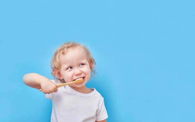 Child brushes his teeth with a wooden bamboo toothbrush on a blue background with a copy space.