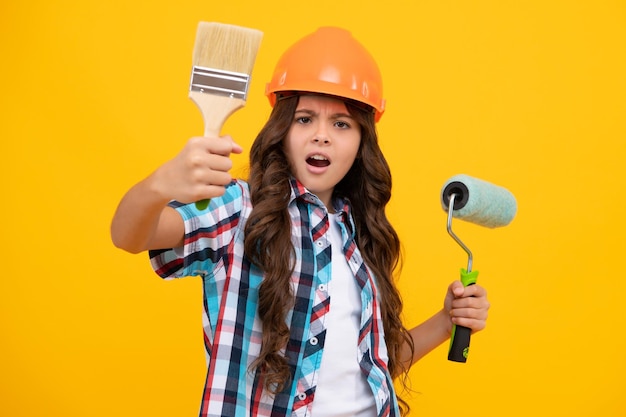 Child builder in hard hat helmet Teenage girl painter with painting brush tool or paint roller Teenager worker isolated on yellow background Kids renovation concept Unhappy upset teenager girl