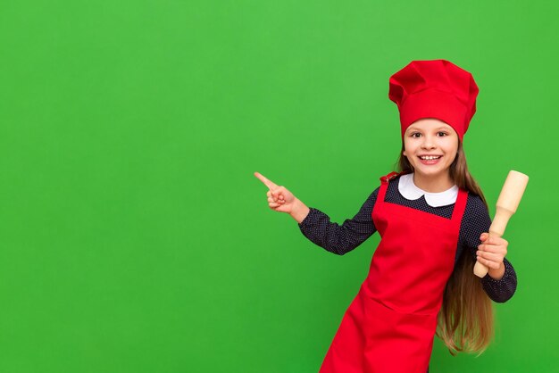 The child chef points to your advertisement on an isolated green background A little girl dressed in a red apron and a cap holds a jump rope for the dough Copy space