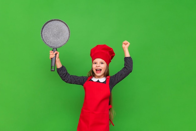 A child in a chef 's costume enthusiastically raises a pancake pan and wide A little girl in a red apron and a hat on a green isolated background