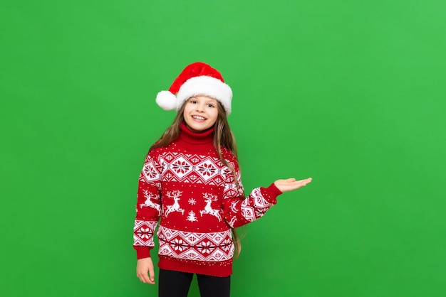 A child in Christmas clothes A beautiful little girl with long hair in a reindeer sweater and a Santa Claus hat holds your advertisement in the palm of her hand on a green isolated background