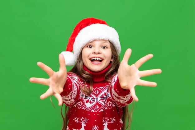 A child in Christmas clothes A beautiful little girl with long hair in a reindeer sweater and Santa Claus hats stretches her arms forward and smiles broadly on a green isolated background