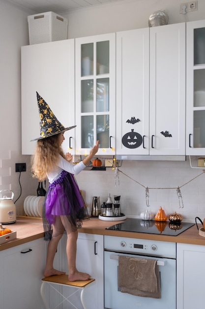 Child decorates the kitchen in home for Halloween Girl in a witch costume plays with the decor for the holiday bats jack lantern pumpkins Autumn comfort in house Scandistyle kitchen loft