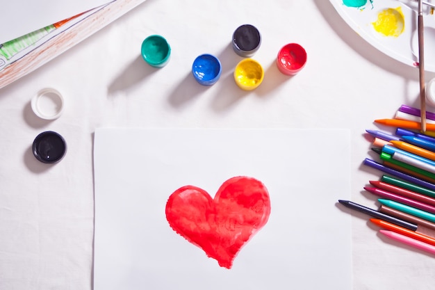 A child drawing red heart with colored paints on the paper.