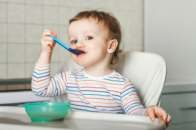 The child eats and holds spoon with fruit, vegetables puree, healthy food for child