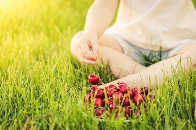 The child eats ripe cherries from a plate, sitting on the green grass on a Sunny day