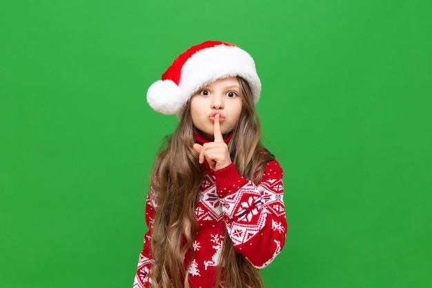 The child gestures that silence should be observed A little girl in a Christmas sweater and Santa Claus hat on a green isolated background