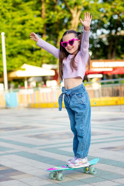 A child girl in an amusement park rides a skateboard in the summer and smiles with happiness near the carousels in sunglasses the concept of summer holidays and school holidays