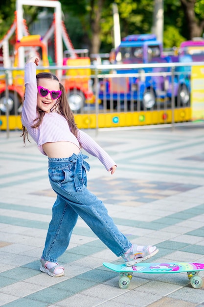 A child girl in an amusement park rides a skateboard in the summer and smiles with happiness near the carousels in sunglasses the concept of summer holidays and school holidays