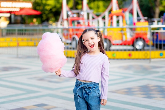 A child girl in an amusement park in the summer eats cotton candy near the carousels the concept of summer holidays and school holidays