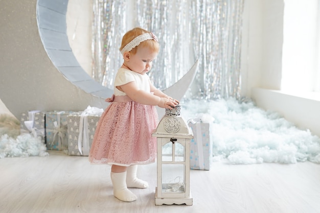 Child girl in beautiful dress holding flashlight near Christmas decorations and gifts, winter New Year holiday