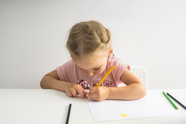 The child girl draws with pencils sitting at a white table on a white background