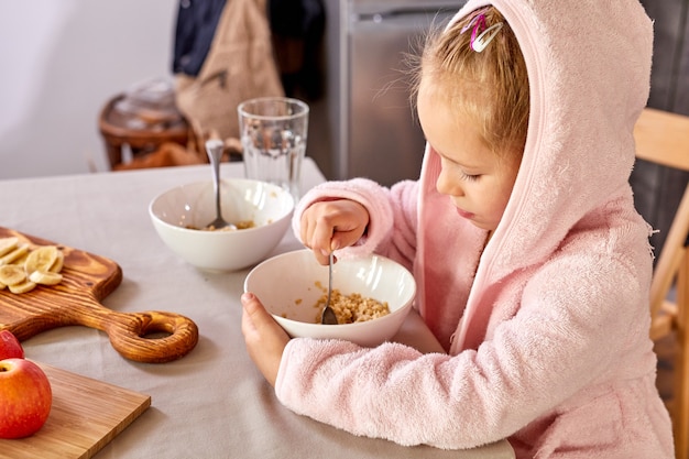 Child girl eating breakfast alone in the kitchen, sit having meal by spoon, wearing domestic clothes