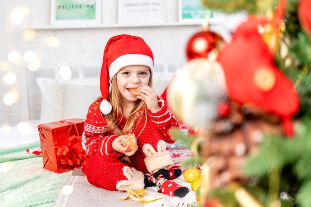 A child girl is on the bed at home eating tangerines and waiting for the new year or Christmas in a red Santa Claus hat and smiling with happiness