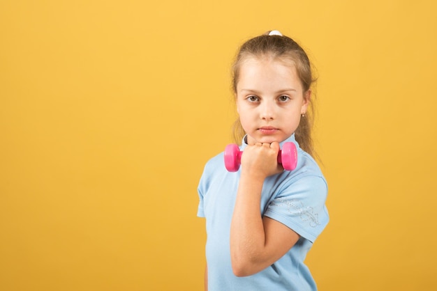 Child girl is doing exercises with dumbbells on yellow studio background