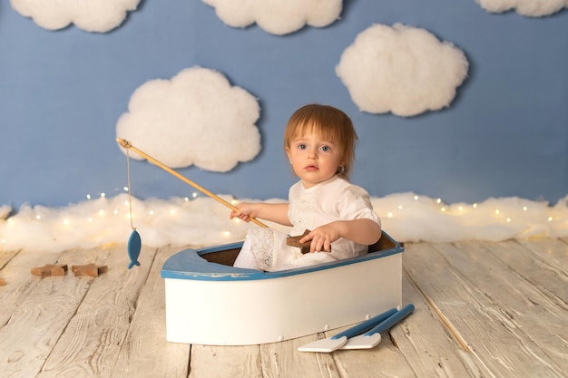 A child girl is sitting in a boat with a fishing rod and a fish against a background of white clouds