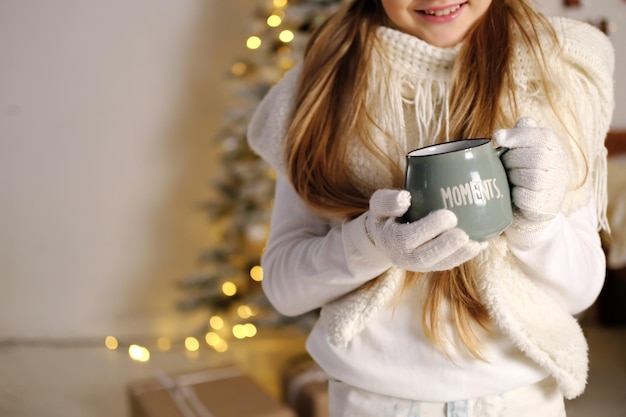 Child girl in a white knitted sweater with a cup of hot drink in her hands poses against the background of Christmas evening lights