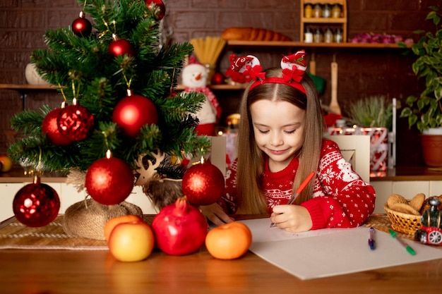 A child girl writes a letter to Santa Claus in a dark kitchen under a Christmas tree with red balls the concept of new year and Christmas