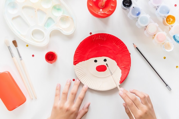 The child glues the parts Santa made from paper plate. Handmade. Project of children's creativity, handicrafts, crafts for kids. Preparation for christmas. Decoration.