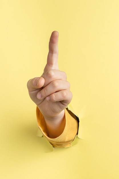 Child hand shows the forefinger up through hole in yellow paper with torn edges concept press the button first