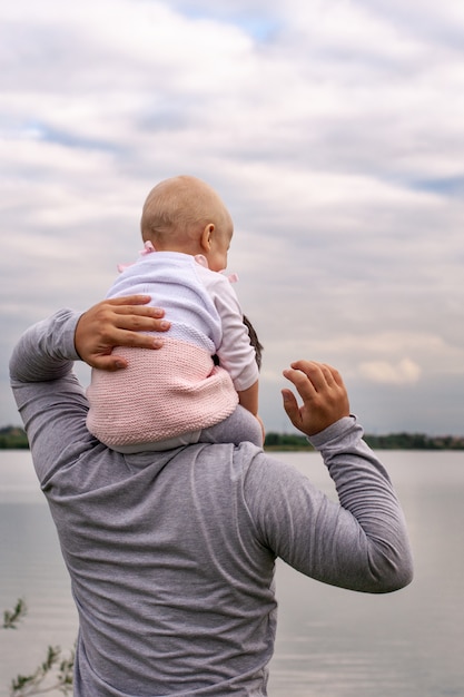 A child on his father neck. Walk near the water. Baby and dad against the sky.