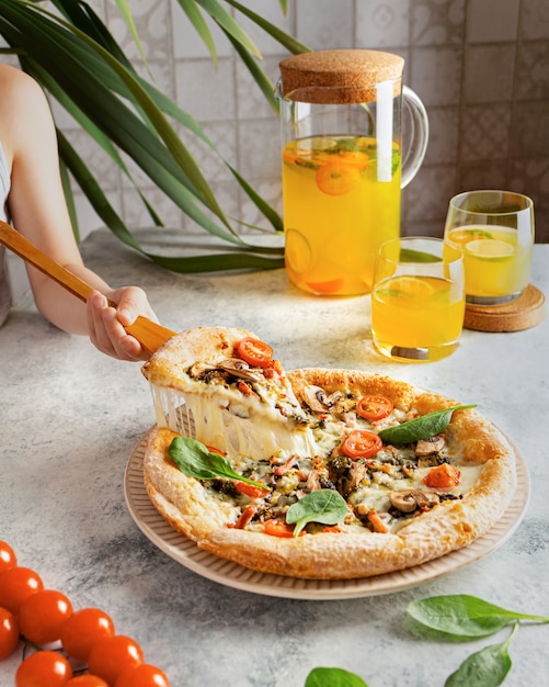 A child holds a hot pizza with melted cheese. A got pizza with melted cheese on a plate on a gray table with lemonade, citrus juice in a glass decanter. Spinach leaves and tomatoes on hot pizza.