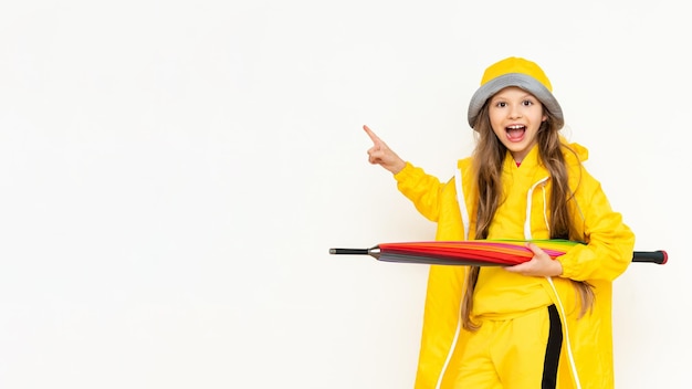 The child holds the umbrella horizontally smiling broadly and pointing at your advertisement A little girl in a yellow raincoat and a panama hat on a white isolated background Copy Space