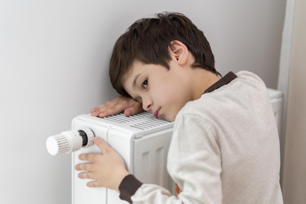 The child hugs the radiator of the home heater with his hands to keep warm