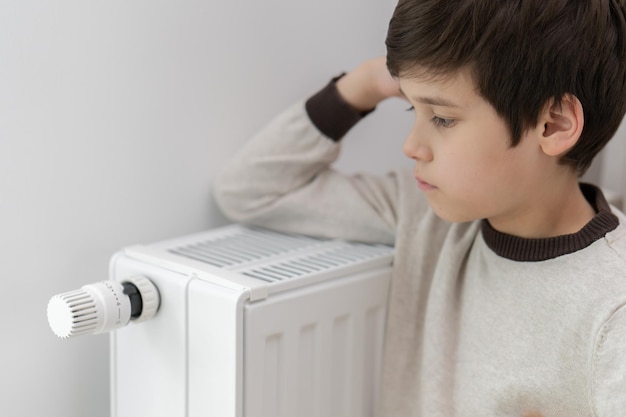 The child hugs the radiator of the home heater with his hands to keep warm