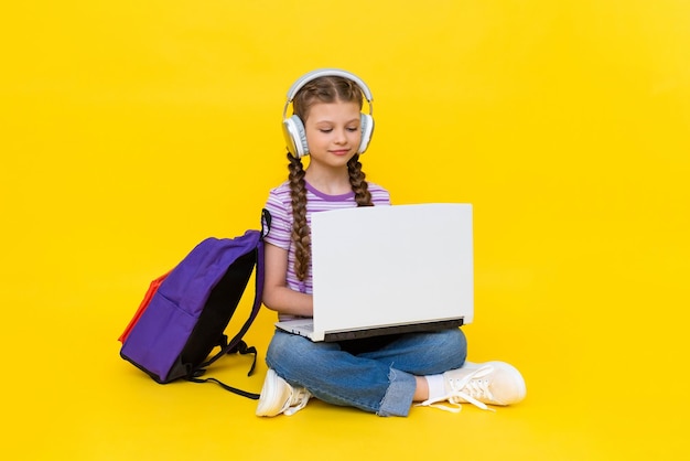 The child is engaged in an online lesson with a laptop A young girl is sitting crosslegged on the floor with a backpack and headphones Yellow isolated background