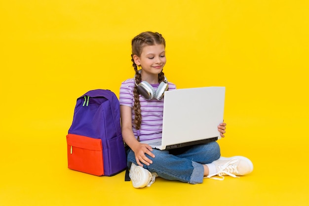 The child is engaged in an online lesson with a laptop A young girl is sitting crosslegged on the floor with a backpack and headphones Yellow isolated background