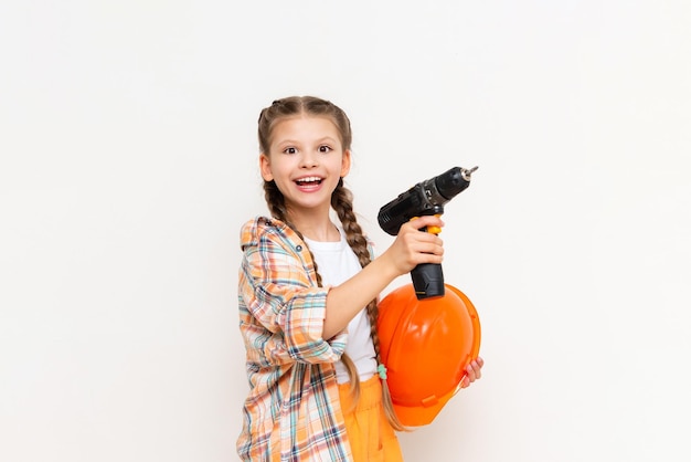 The child is holding a cordless drill and a protective construction helmet on a white insulated background A little girl with long pigtails holding a screwdriver is smiling broadly Copy Space