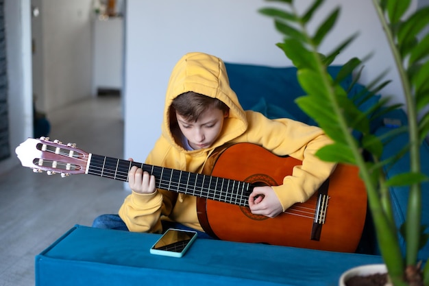 Child is practicing acoustic guitar in his room Cute boy playing acoustic guitar dressed casual jeans and yellow sweatshirt on the cozy sofa at home Concept of hobbies