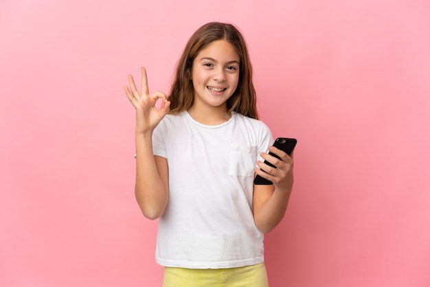 Child over isolated pink background using mobile phone and doing OK sign